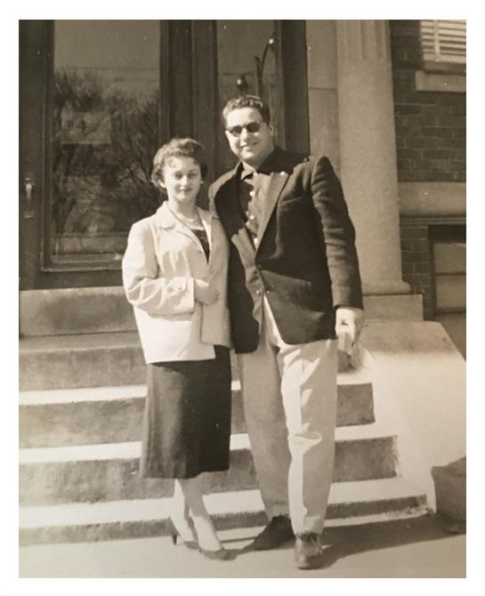 A couple smiles on the steps of a mid-20th century building.