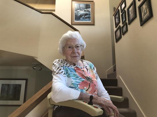 An elderly woman relaxes in a chair near stairs, surrounded by framed photos and a cozy atmosphere.