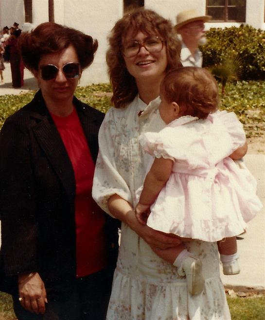 Three women stand together in a sunny park, one holding a small girl while smiling warmly.