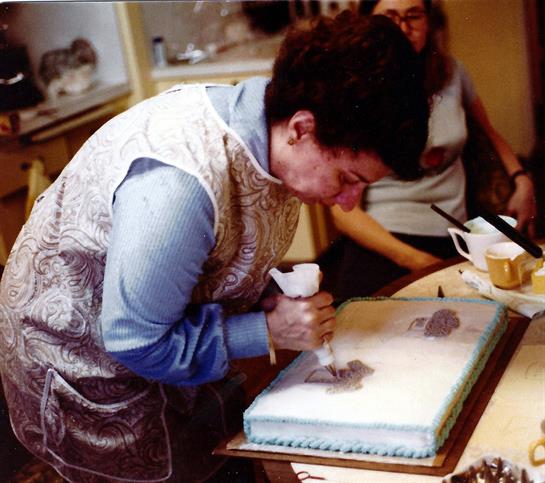 An elderly woman carefully pipes icing onto a cake while friends chat nearby in a warm kitchen.
