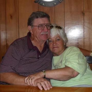 An elderly couple sits closely together at a wooden table, enjoying each other's company and smiles.
