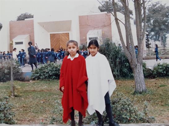 Two girls in red and white ponchos pose outside a school as classmates observe the event.