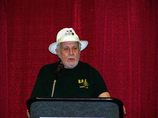 A man at a podium shares knowledge with an audience, wearing a pin-adorned hat.