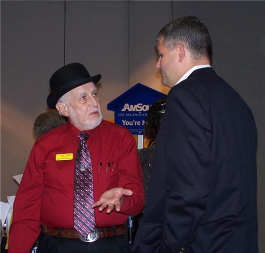 Two men chat in a conference room; one in a red shirt and hat, the other in formal wear.