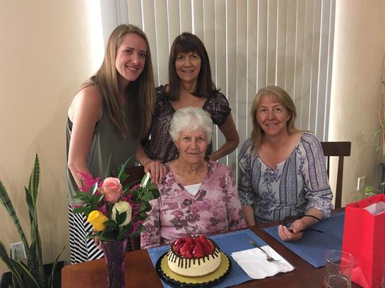 Four generations unite in a warm celebration with cake and flowers for a special birthday.