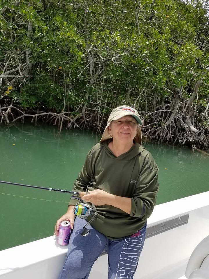 A woman sits on a boat, fishing in calm waters surrounded by lush mangroves under bright sunlight.