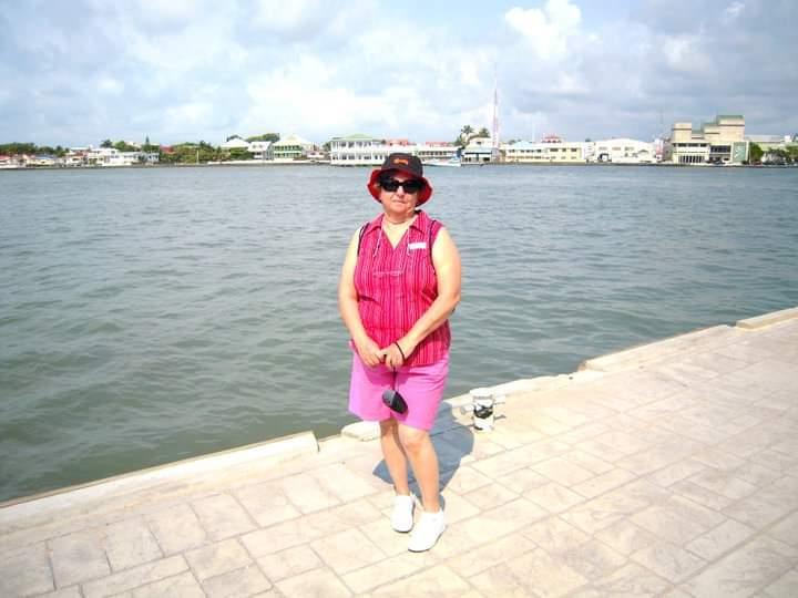 A woman stands smiling by the water, dressed in a bright pink outfit, enjoying a sunny day.