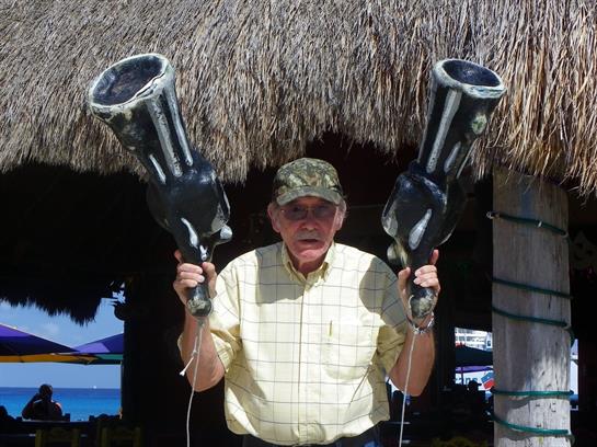 Man in a cap poses with large novelty guns under a thatched roof, showcasing a playful spirit.