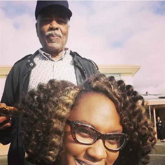 Woman with curly hair and glasses smiles while older man stands behind her, holding food.