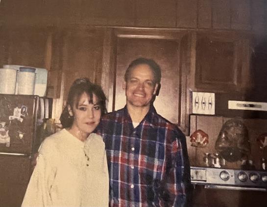 A couple smiles while standing together in a cozy kitchen filled with wooden cabinets.