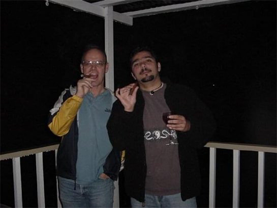 Two men relax on a balcony at night, smoking cigars and holding drinks while chatting together.