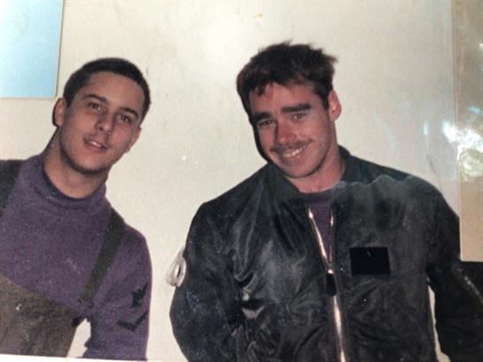 Two young men stand close together, wearing military jackets, smiling happily indoors.