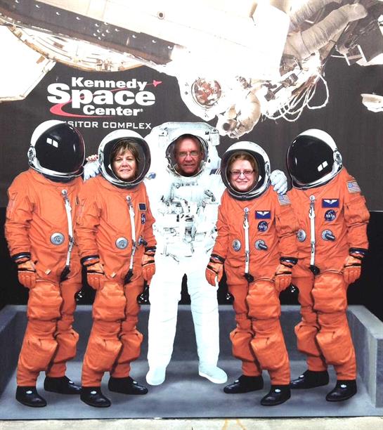 Four people wearing bright orange astronaut suits pose together at a space center.