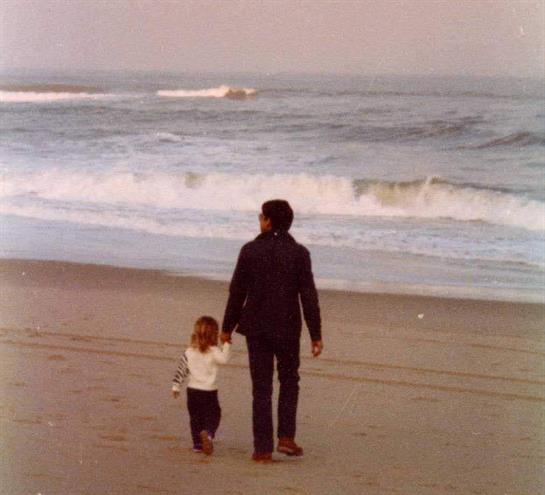 Adult and child stroll together along the sandy beach, waves gently crashing in the background.