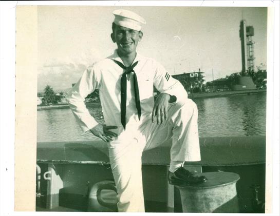 A young sailor in white uniform stands confidently with one foot on a boat, smiling.