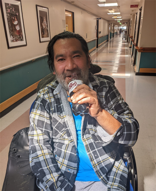 A man with a beard smiles as he holds a can while seated in a wheelchair in a hallway.