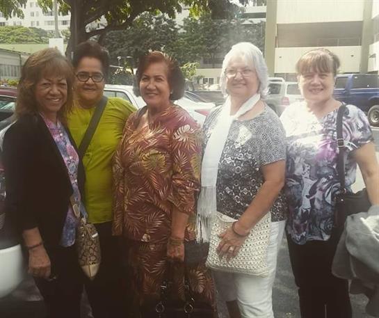 Six women stand together, smiling warmly, enjoying a pleasant day in an urban location.