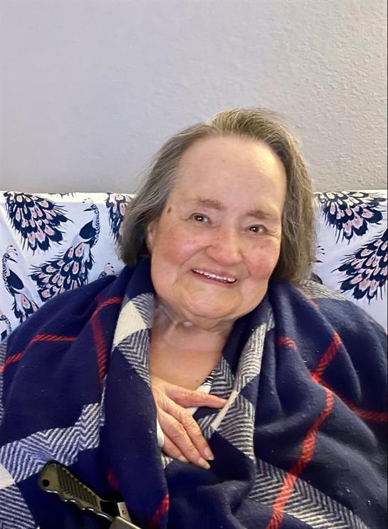 Elder woman smiles warmly while sitting on a couch, wrapped in a blanket, enjoying a moment at home.