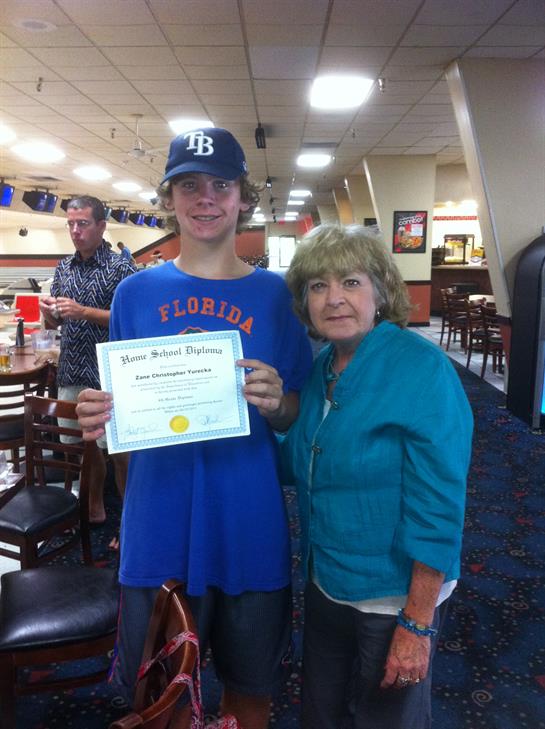 A young bowler stands proudly with his certificate next to a supportive woman at a bowling alley.