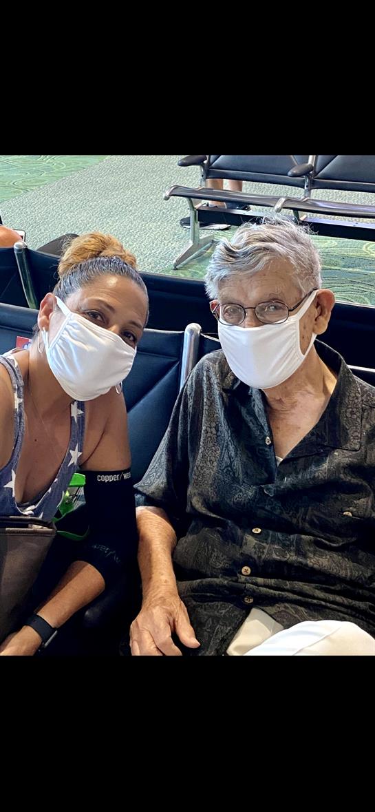 Two individuals are smiling while seated in an airport, both wearing masks and waiting.