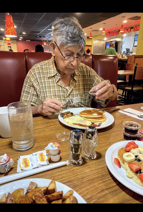 An elderly man carefully eats a breakfast of pancakes and sausages in a bustling diner setting.