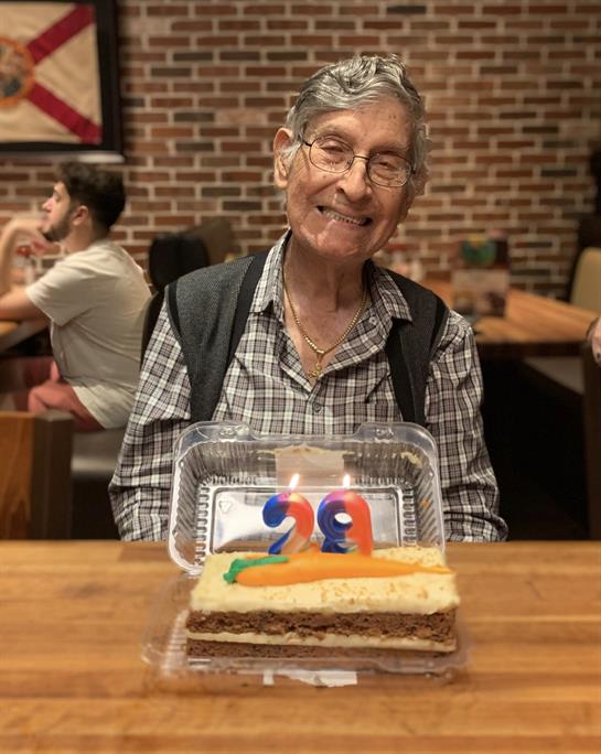 A cheerful elderly man smiles widely while holding a birthday cake with candles in a cafe.