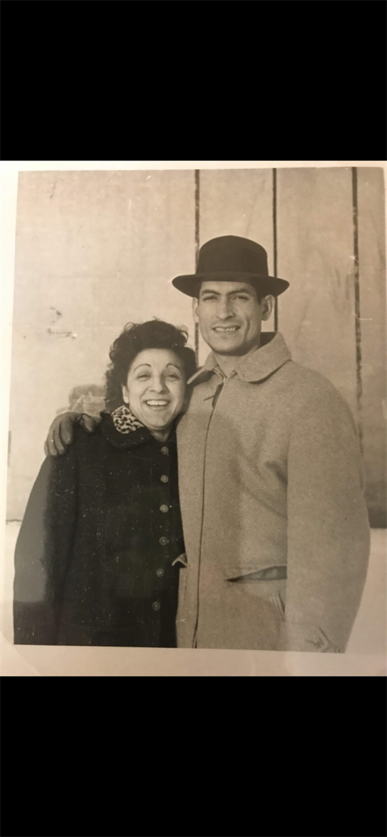 A joyful couple stands close together, sharing smiles in winter attire against a rustic backdrop.