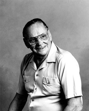An older man smiles warmly, sitting comfortably against a simple backdrop.