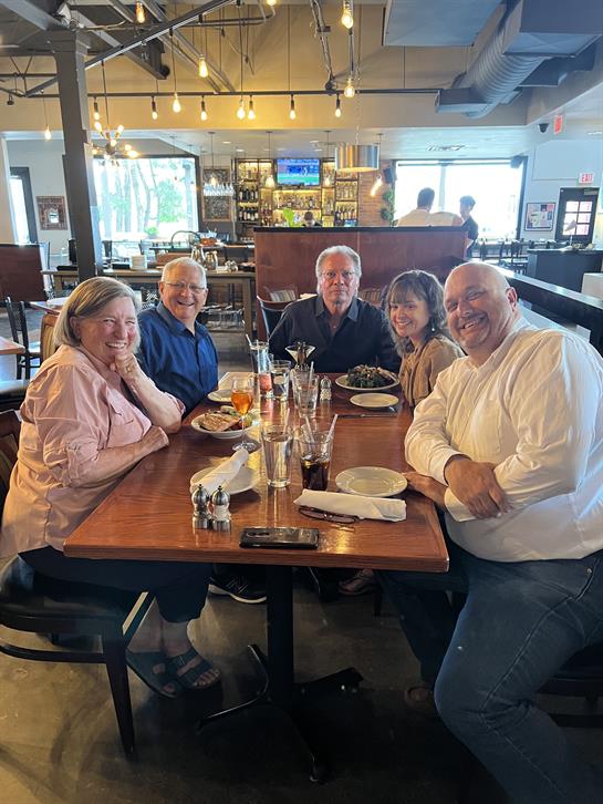 A group of five friends gathers around a table sharing food and drinks while smiling.