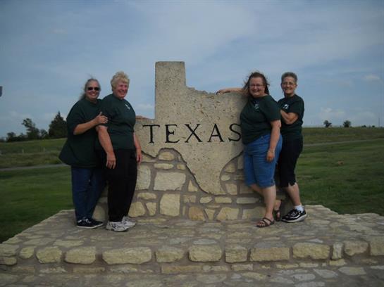 Four friends pose joyfully at the Texas state border, showcasing unity and happiness outdoors.