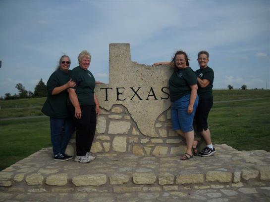 Friends happily gather around Texas state sign at roadside park, enjoying a sunny day together.