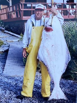 A fisherman shows off a large halibut he caught, standing by a rustic dock surrounded by nature.