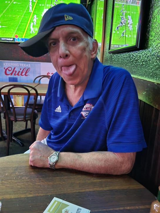 An older man in a blue shirt sits at a sports bar, playfully sticking out his tongue.