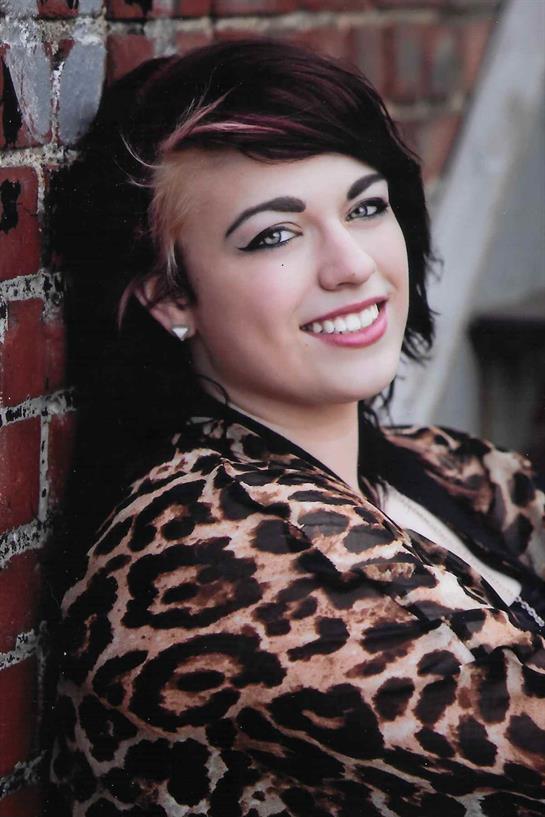 A woman smiles warmly while leaning against a brick wall, showcasing her fashionable style.