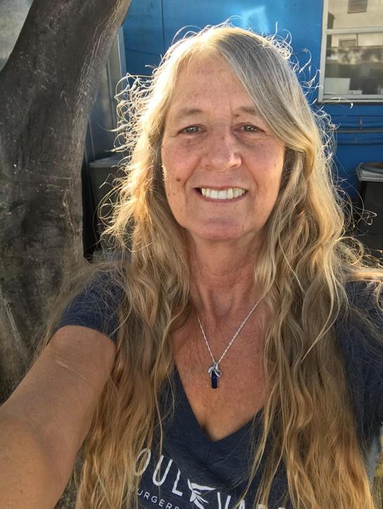 A woman with long hair smiles warmly in a sunlit outdoor setting.