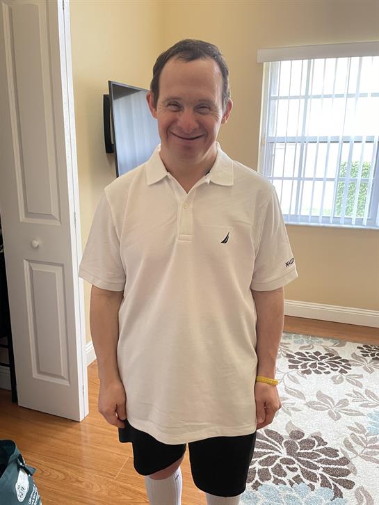 A man with a cheerful expression stands indoors, dressed in a white polo shirt, smiling happily.