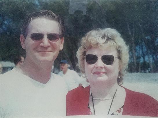 A couple smiles with sunglasses on, enjoying a sunny day at a beach, surrounded by friends.