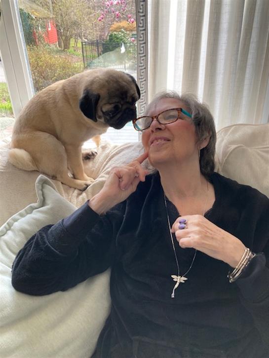 A woman enjoys a light-hearted moment with her pug dog on a soft sofa, smiling and relaxed indoors.