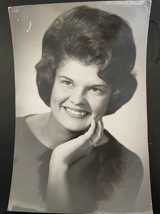 A young woman with wavy hair smiles warmly while resting her chin on her hand in a studio setup.