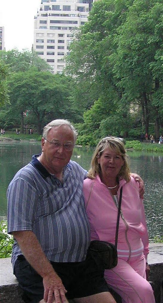 Two people stand close together by a tranquil lake, surrounded by lush trees and nature.