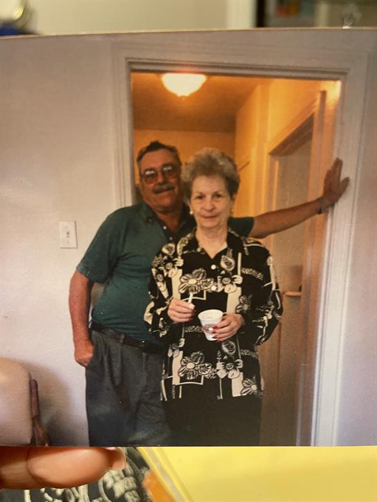 Elderly couple stands in their doorway, sharing smiles and holding cups of tea in a warm atmosphere.