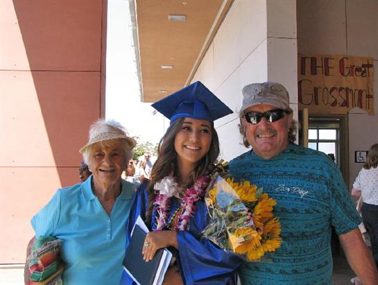 A young woman in a graduation gown holds flowers while posing with her family outdoors.