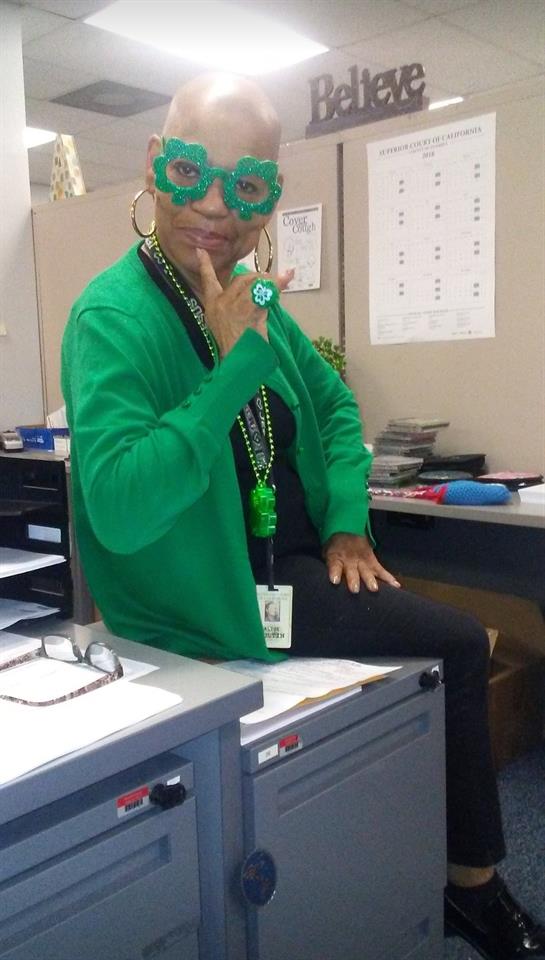 A woman in vibrant green clothing strikes a playful pose at her workplace, celebrating a holiday.