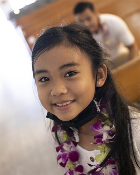 A joyful girl with a flower lei beams at the camera in a lively indoor environment.