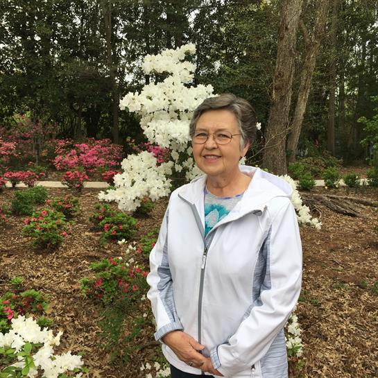 A woman stands happily in a garden filled with vibrant azalea flowers under a clear sky.