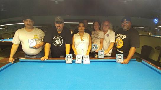 Friends stand behind a blue pool table, proudly displaying trophies after a tournament.