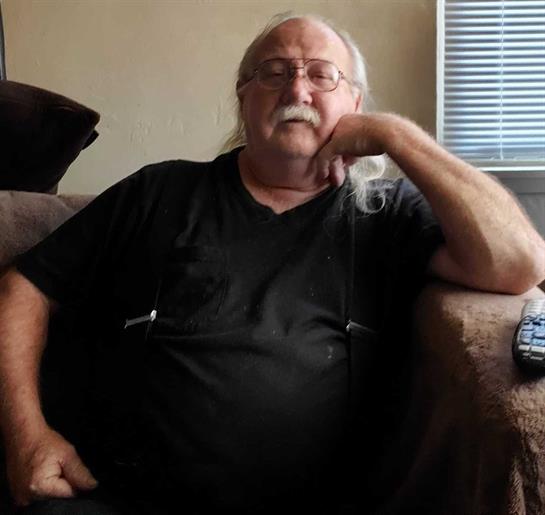 An older man dressed in black relaxes on a couch, appearing thoughtful and comfortable in his home.
