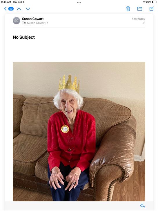 An elderly woman in a red outfit smiles brightly while sitting on a couch with a crown.