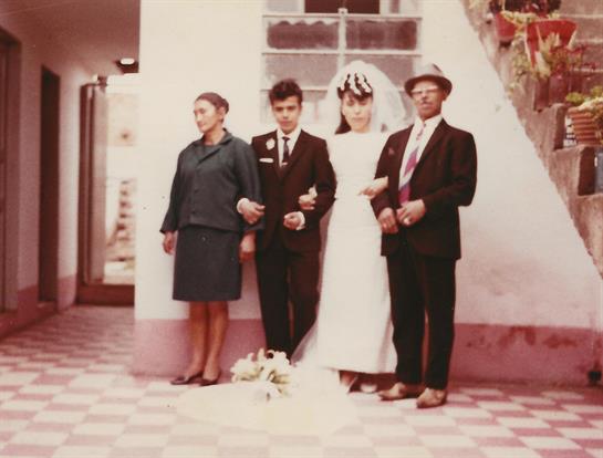 A newly married couple stands proudly with their parents in a bright indoor location.