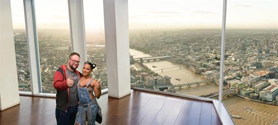 A couple poses happily at a high-rise window, admiring the stunning skyline and river below.